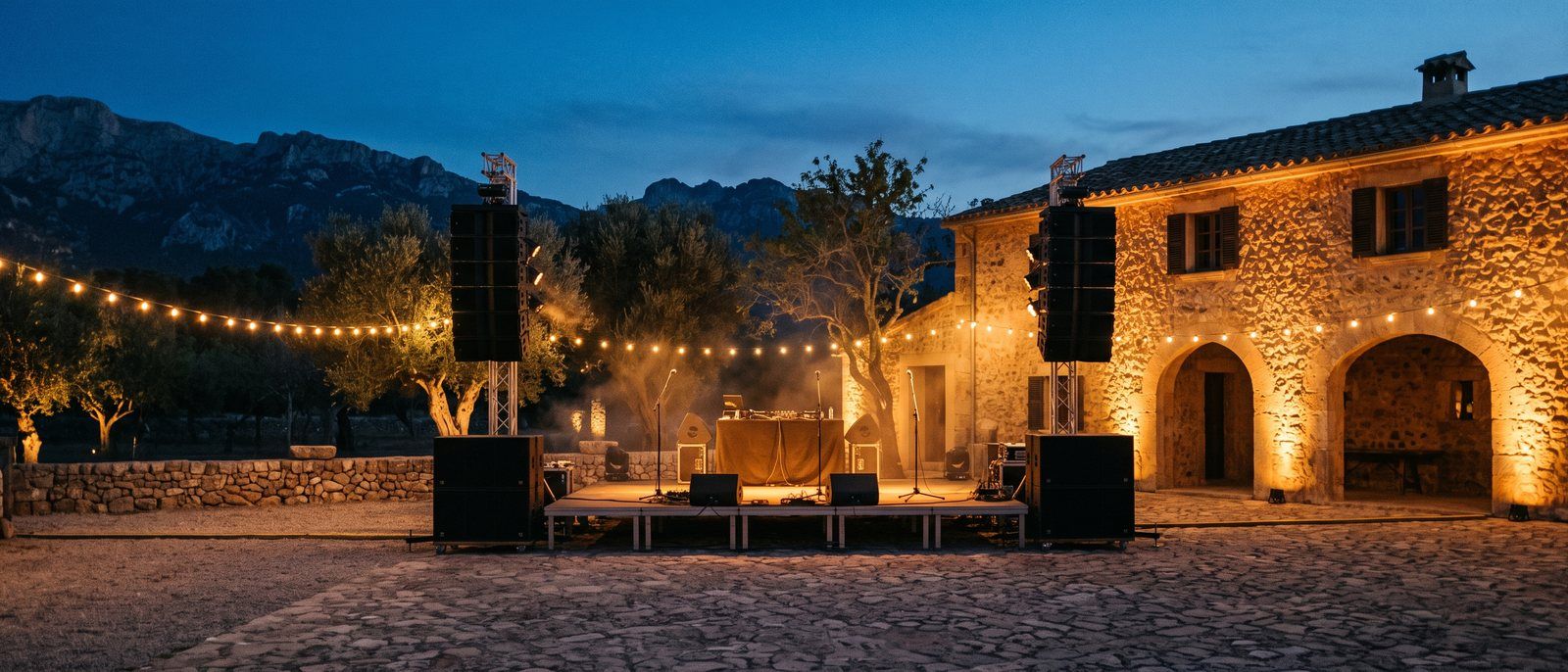 Cobbled courtyard of a Mallorcan finca at dusk with two sound stacks flanking a wooden stage