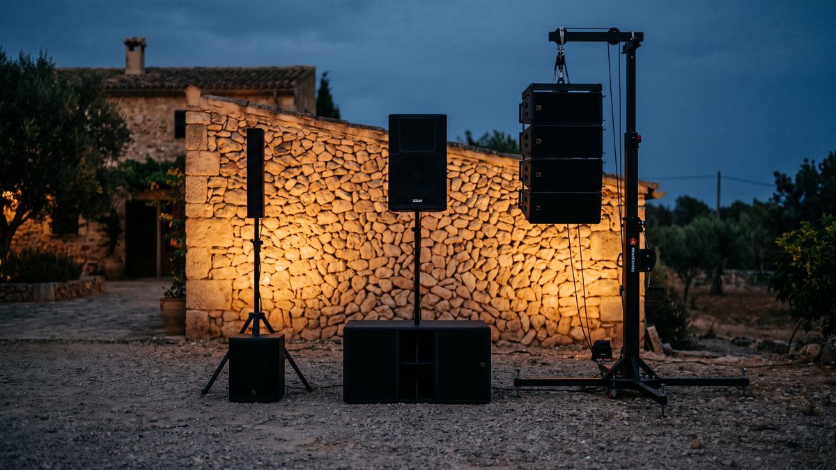 Trois tailles de système PA alignées sur du gravier devant un mur en pierre majorquin : colonne portable, stack enceinte sur subwoofer et line array suspendu à une tour élévatrice