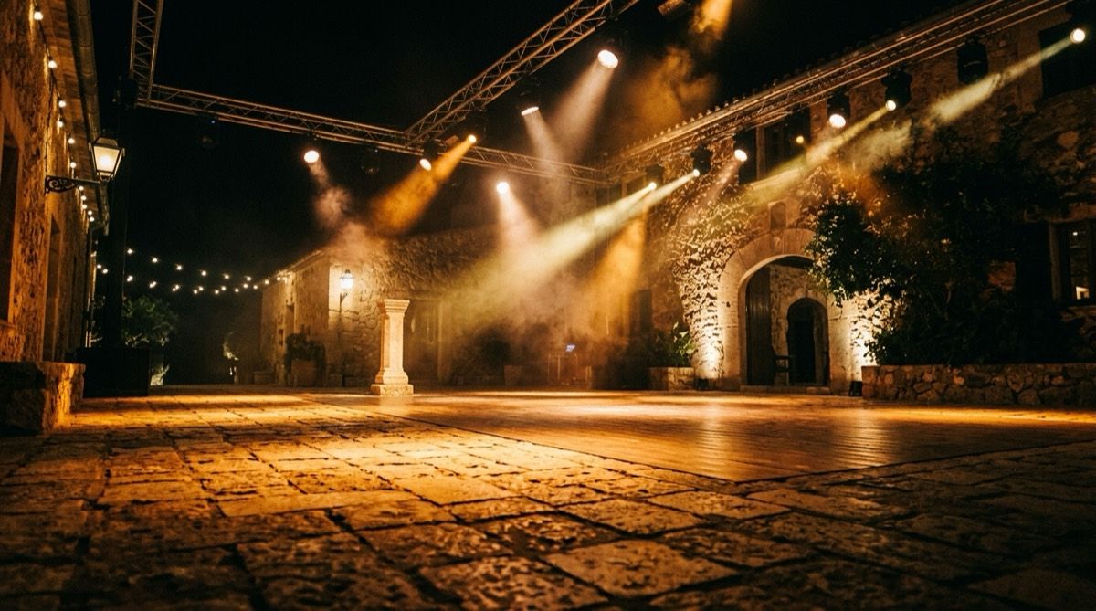 Pista de ball a l'aire lliure en una possessió banyada per focus wash LED en ambre, amb caps mòbils a dalt i fum travessant els feixos